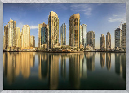 A panoramic photograph of the Dubai Marina skyline with its golden skyscrapers reflecting in the calm, dark water below, all under a clear blue sky and framed in silver. Wall Art