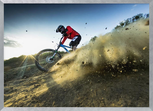 A mountain biker wearing a red jersey and helmet speeds down a steep, dusty trail, kicking up a large cloud of dirt. The sun is setting in the background, creating a lens flare against a clear blue sky. The image is captured from a low angle, emphasizing the action and speed, and is presented within a silver frame. Artwork