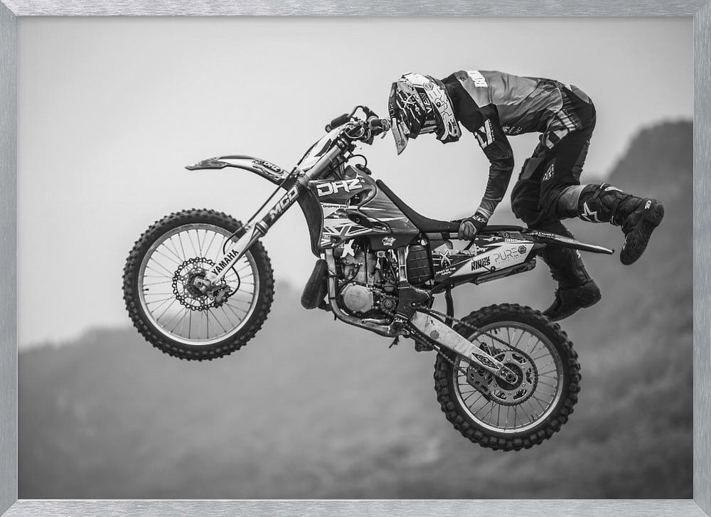A dynamic black and white photograph of a motocross rider in mid-air, performing a jump. The rider, wearing full protective gear and a helmet, is leaning forward over the handlebars of the dirt bike. The bike is suspended against a blurry, light gray background, emphasizing the action and height of the jump. The image is enclosed in a silver frame. Wall Art