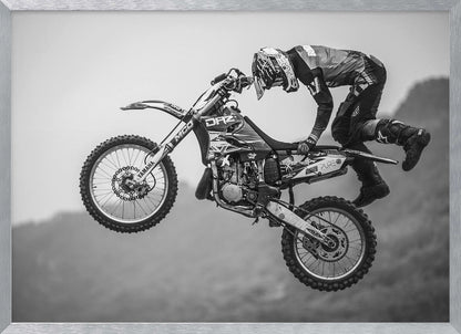 A dynamic black and white photograph of a motocross rider in mid-air, performing a jump. The rider, wearing full protective gear and a helmet, is leaning forward over the handlebars of the dirt bike. The bike is suspended against a blurry, light gray background, emphasizing the action and height of the jump. The image is enclosed in a silver frame. Wall Art