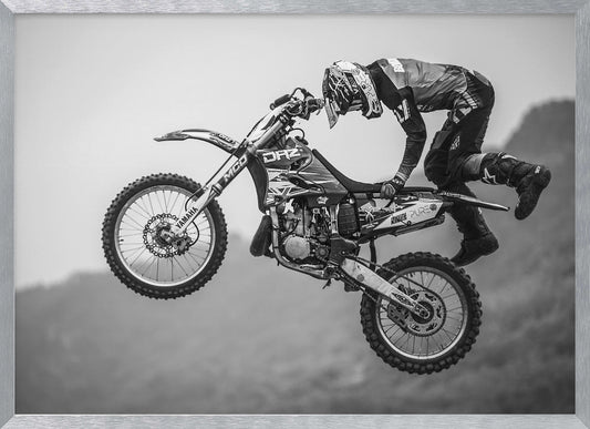 A dynamic black and white photograph of a motocross rider in mid-air, performing a jump. The rider, wearing full protective gear and a helmet, is leaning forward over the handlebars of the dirt bike. The bike is suspended against a blurry, light gray background, emphasizing the action and height of the jump. The image is enclosed in a silver frame. Wall Art