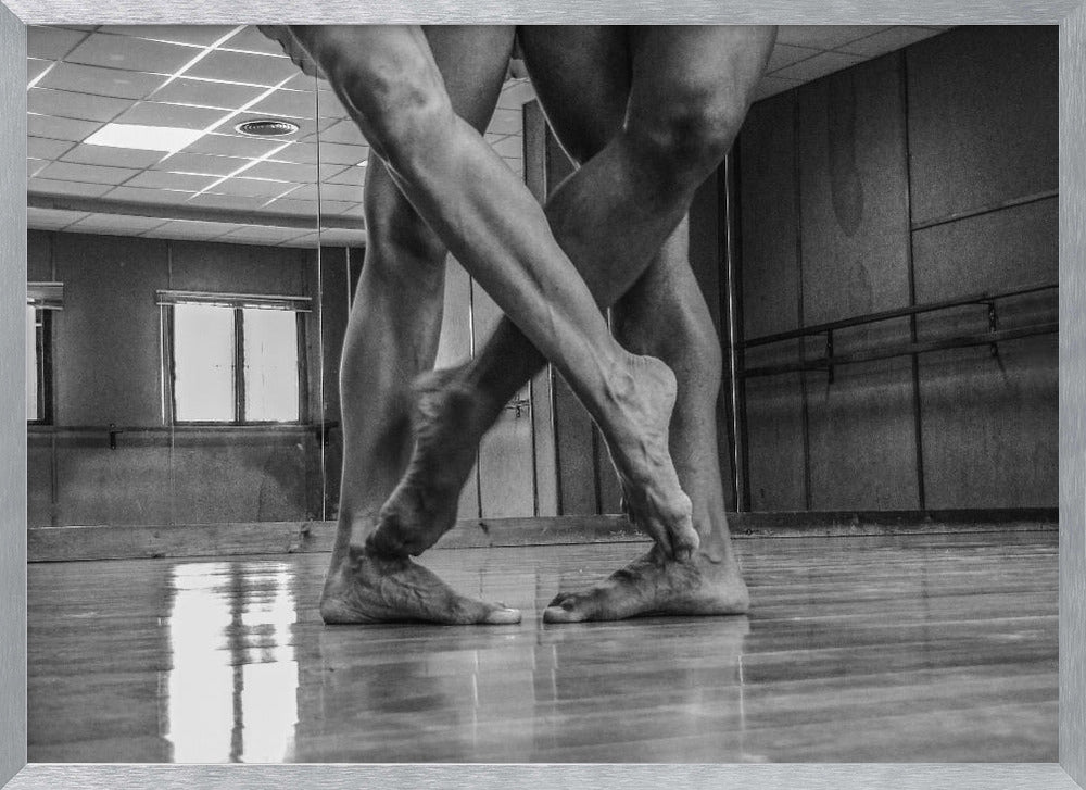 A dramatic low-angle, black and white photograph capturing the muscular legs and bare feet of two dancers intertwined in a dance studio. Their limbs cross in a display of strength and grace on a reflective wooden floor, with the studio's mirrored wall and barre visible in the background. The image is enclosed in a silver-grey frame. Decor