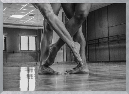 A dramatic low-angle, black and white photograph capturing the muscular legs and bare feet of two dancers intertwined in a dance studio. Their limbs cross in a display of strength and grace on a reflective wooden floor, with the studio's mirrored wall and barre visible in the background. The image is enclosed in a silver-grey frame. Decor