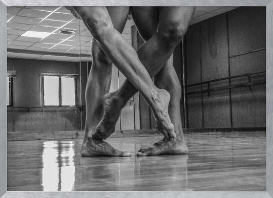 A dramatic low-angle, black and white photograph capturing the muscular legs and bare feet of two dancers intertwined in a dance studio. Their limbs cross in a display of strength and grace on a reflective wooden floor, with the studio's mirrored wall and barre visible in the background. The image is enclosed in a silver-grey frame. Decor