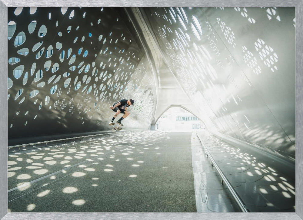 A skateboarder is captured mid-air performing a trick inside a futuristic, metallic tunnel with patterned cutouts that cast dappled light and shadows across the scene, all enclosed within a silver frame. Artwork