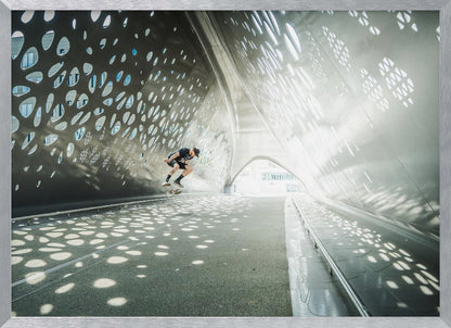 A skateboarder is captured mid-air performing a trick inside a futuristic, metallic tunnel with patterned cutouts that cast dappled light and shadows across the scene, all enclosed within a silver frame. Artwork