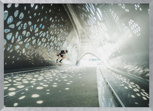 A skateboarder is captured mid-air performing a trick inside a futuristic, metallic tunnel with patterned cutouts that cast dappled light and shadows across the scene, all enclosed within a silver frame. Artwork