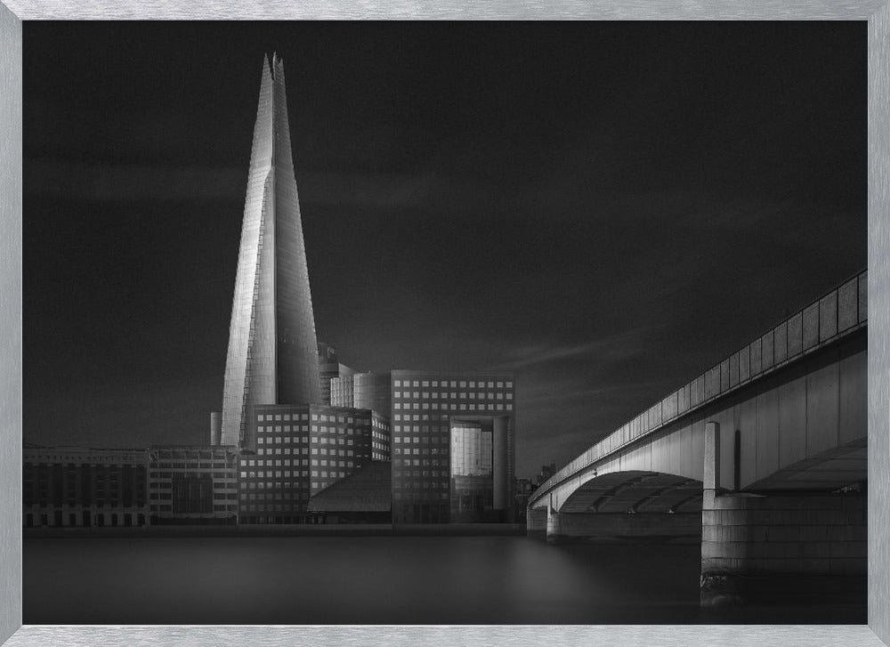 A dramatic black and white photograph of the London skyline featuring The Shard skyscraper and London Bridge. The image is shot at dusk or night, with The Shard brightly lit against a dark, moody sky. The River Thames in the foreground is calm and reflective, captured with a long exposure. Artwork