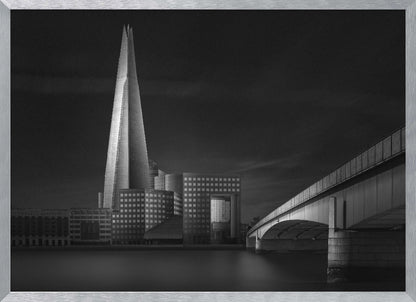 A dramatic black and white photograph of the London skyline featuring The Shard skyscraper and London Bridge. The image is shot at dusk or night, with The Shard brightly lit against a dark, moody sky. The River Thames in the foreground is calm and reflective, captured with a long exposure. Artwork