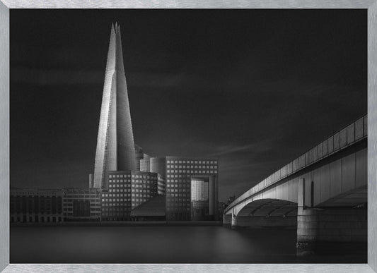 A dramatic black and white photograph of the London skyline featuring The Shard skyscraper and London Bridge. The image is shot at dusk or night, with The Shard brightly lit against a dark, moody sky. The River Thames in the foreground is calm and reflective, captured with a long exposure. Artwork