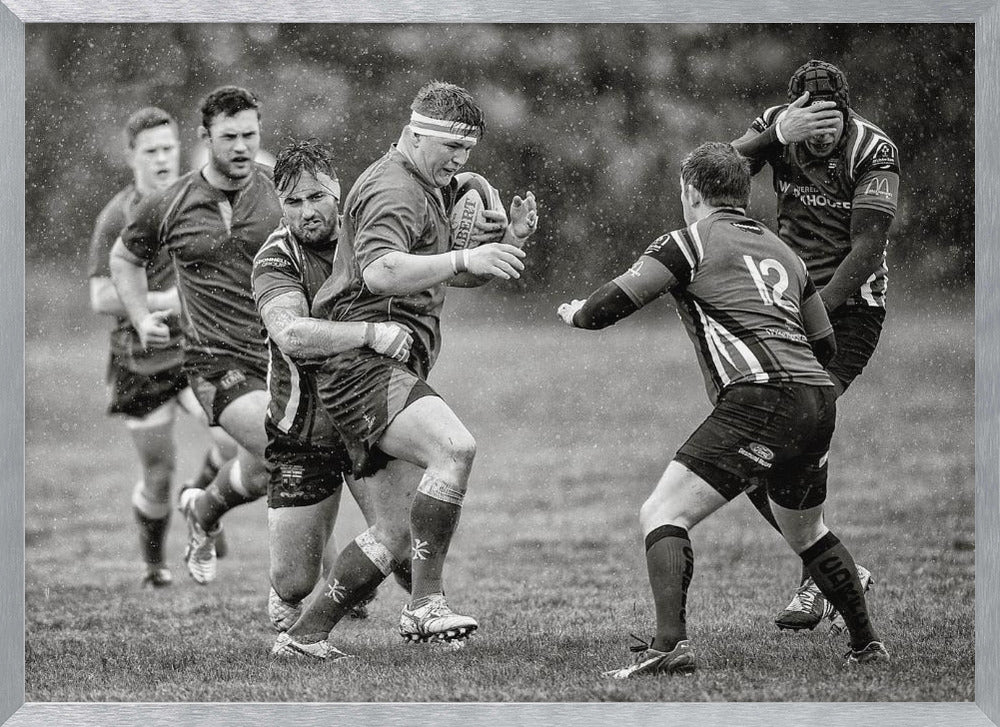 A dynamic black and white photograph of a rugby match being played in the rain. One player charges forward with the ball while an opponent wraps his arms around him in a tackle. Other players are in motion around them, capturing the intensity and physicality of the sport. Decor
