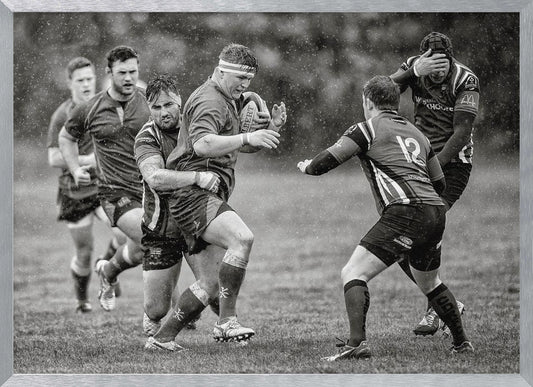 A dynamic black and white photograph of a rugby match being played in the rain. One player charges forward with the ball while an opponent wraps his arms around him in a tackle. Other players are in motion around them, capturing the intensity and physicality of the sport. Decor