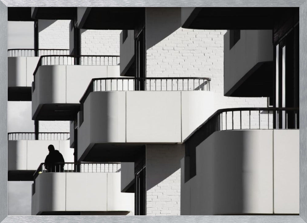 A black and white architectural photograph of a modern building with repeating geometric balconies. Strong sunlight creates deep shadows, emphasizing the lines and curves of the structure. A lone silhouetted figure stands on one of the lower balconies. Decor