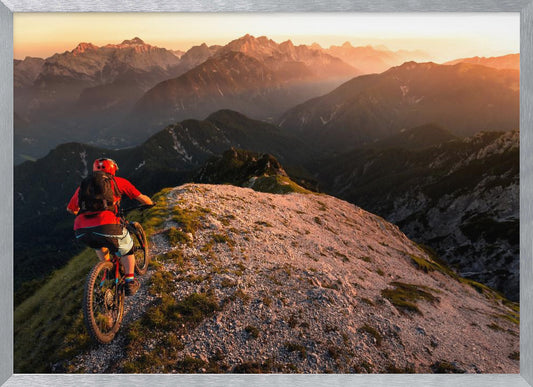 A mountain biker wearing a red shirt and helmet rides along a narrow, rocky mountain ridge at sunset. The background features a vast landscape of layered mountain ranges glowing in the warm, golden light of the setting sun. Poster