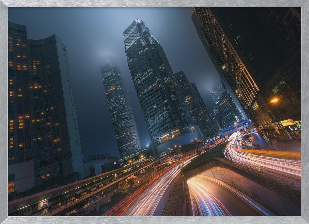A framed, low-angle photograph capturing a bustling city at night, with towering skyscrapers shrouded in fog. Long-exposure creates vibrant streaks of light from traffic on a multi-level highway, illuminating the dark, modern urban landscape. Wall Art