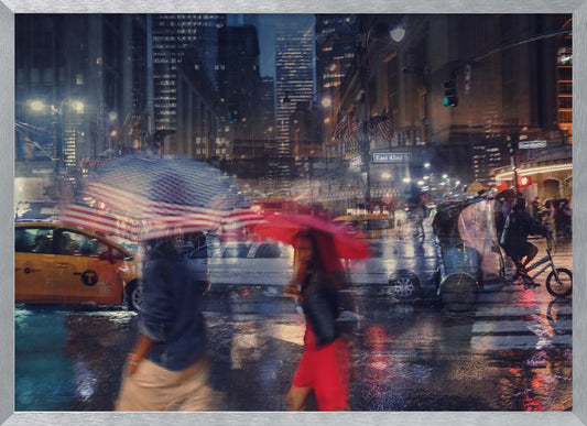 An artistic motion-blur photograph of a rainy night on East 42nd Street in New York City. Pedestrians with umbrellas, one with an American flag design and another bright red, cross the wet street as city lights and a yellow taxi reflect on the pavement, creating a vibrant, impressionistic scene within a silver frame. Decor