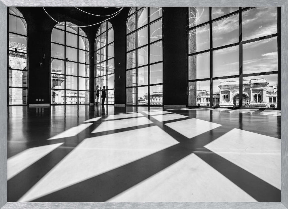 A striking black and white photograph of a spacious, modern hall with a polished floor. Dramatic shadows from large, arched windows are cast across the floor. Through the windows, a view of a classical European city with a triumphal arch can be seen. Print
