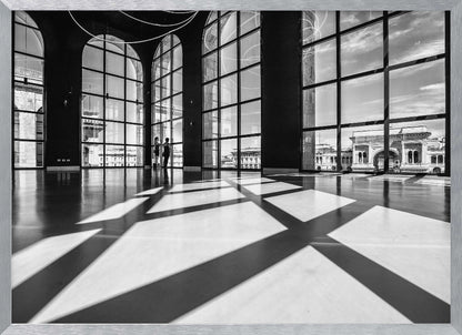 A striking black and white photograph of a spacious, modern hall with a polished floor. Dramatic shadows from large, arched windows are cast across the floor. Through the windows, a view of a classical European city with a triumphal arch can be seen. Print