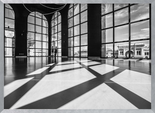 A striking black and white photograph of a spacious, modern hall with a polished floor. Dramatic shadows from large, arched windows are cast across the floor. Through the windows, a view of a classical European city with a triumphal arch can be seen. Print