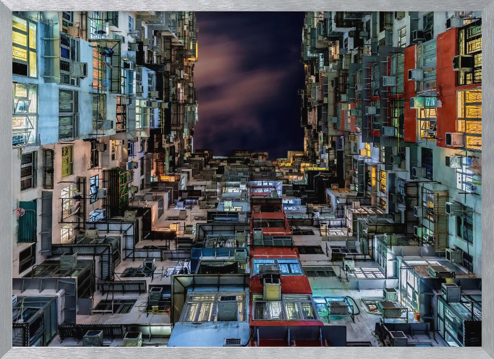 A low-angle nighttime view looking up from a courtyard surrounded by extremely dense, stacked apartment buildings with many windows lit in yellow, blue, and white, revealing a small patch of dark, cloudy sky. The image is presented within a brushed silver frame. Artwork
