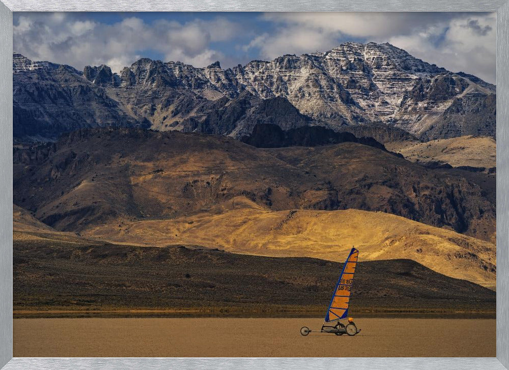 A framed photograph of a land sailor with a bright orange and blue sail on a vast, flat desert floor. In the background, majestic snow-dusted mountains rise against a cloudy blue sky. Artwork