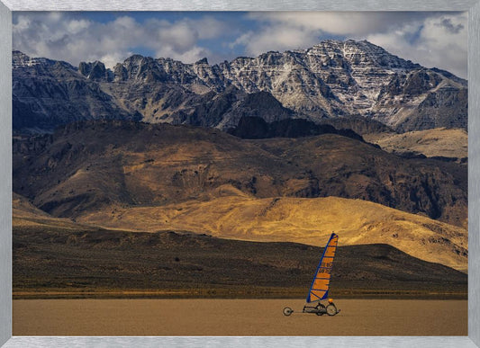 A framed photograph of a land sailor with a bright orange and blue sail on a vast, flat desert floor. In the background, majestic snow-dusted mountains rise against a cloudy blue sky. Artwork