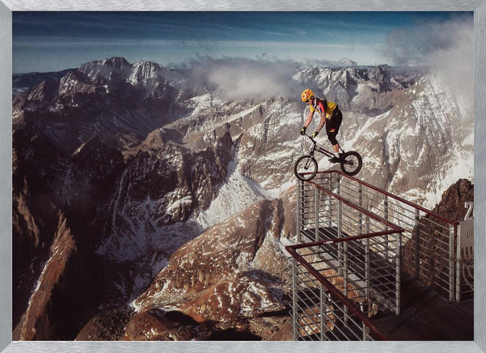 A trials cyclist wearing a yellow helmet balances precariously on the railing of a metal walkway high in the mountains, with a dramatic background of snow-dusted peaks and a blue sky. Poster