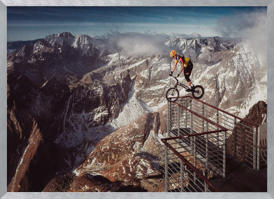 A trials cyclist wearing a yellow helmet balances precariously on the railing of a metal walkway high in the mountains, with a dramatic background of snow-dusted peaks and a blue sky. Poster
