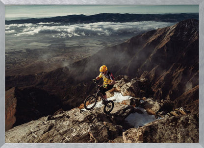 A mountain biker in a yellow helmet and jersey navigates a treacherous, rocky mountain ridge. Below, a vast valley is filled with a sea of clouds, with distant mountain ranges visible on the horizon. Print