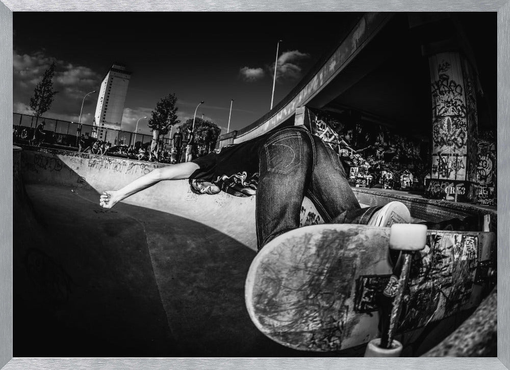 A dynamic, wide-angle black and white photograph of a skateboarder at a skatepark. The skater is bent backwards over the curved edge of a concrete bowl, with one hand touching the surface. Their skateboard is propped up in the foreground, and the background is filled with graffiti-covered walls, spectators, and a tall building against a dark, cloudy sky. Artwork