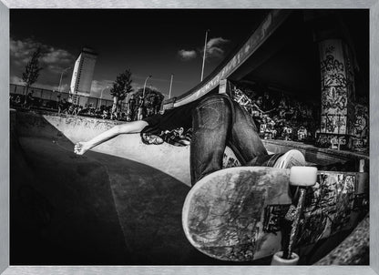 A dynamic, wide-angle black and white photograph of a skateboarder at a skatepark. The skater is bent backwards over the curved edge of a concrete bowl, with one hand touching the surface. Their skateboard is propped up in the foreground, and the background is filled with graffiti-covered walls, spectators, and a tall building against a dark, cloudy sky. Artwork