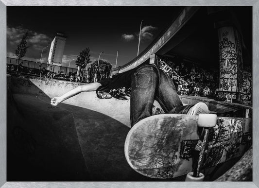 A dynamic, wide-angle black and white photograph of a skateboarder at a skatepark. The skater is bent backwards over the curved edge of a concrete bowl, with one hand touching the surface. Their skateboard is propped up in the foreground, and the background is filled with graffiti-covered walls, spectators, and a tall building against a dark, cloudy sky. Artwork