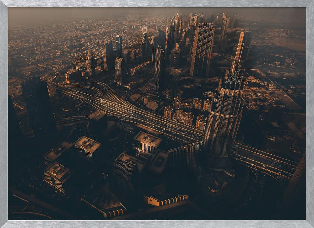 An aerial view of a sprawling modern city at dusk, with the warm glow of sunset casting long shadows over skyscrapers and illuminating a complex network of highways. The photograph has a warm, sepia-like tone and is presented within a silver-colored frame. Decor