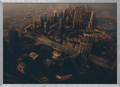 An aerial view of a sprawling modern city at dusk, with the warm glow of sunset casting long shadows over skyscrapers and illuminating a complex network of highways. The photograph has a warm, sepia-like tone and is presented within a silver-colored frame. Decor