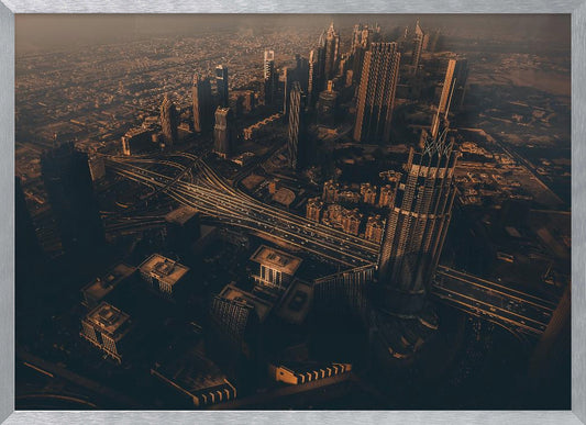 An aerial view of a sprawling modern city at dusk, with the warm glow of sunset casting long shadows over skyscrapers and illuminating a complex network of highways. The photograph has a warm, sepia-like tone and is presented within a silver-colored frame. Decor