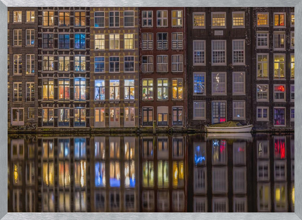 A framed nighttime photograph of a row of traditional Amsterdam canal houses with many illuminated windows, their colorful lights reflecting perfectly in the calm canal water below. A small white boat is moored at the water's edge. Decor