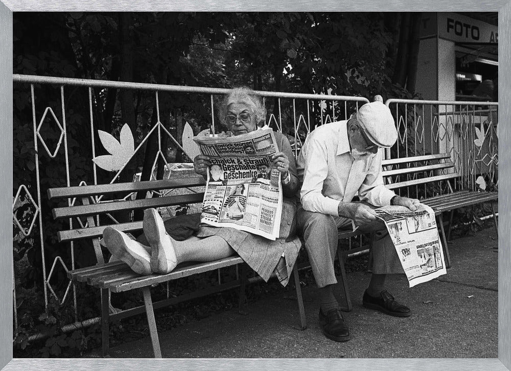 A framed black and white photograph of an elderly couple sitting on a park bench, both deeply absorbed in reading their newspapers. The woman on the left has her feet propped up on the bench, while the man on the right sits next to her, looking down at his paper. Poster
