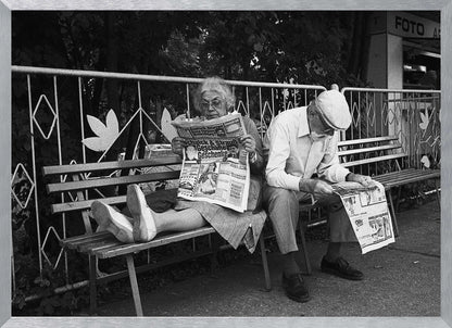 A framed black and white photograph of an elderly couple sitting on a park bench, both deeply absorbed in reading their newspapers. The woman on the left has her feet propped up on the bench, while the man on the right sits next to her, looking down at his paper. Poster