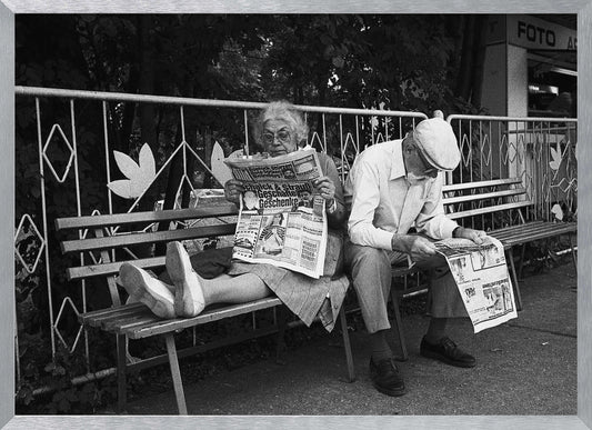 A framed black and white photograph of an elderly couple sitting on a park bench, both deeply absorbed in reading their newspapers. The woman on the left has her feet propped up on the bench, while the man on the right sits next to her, looking down at his paper. Poster