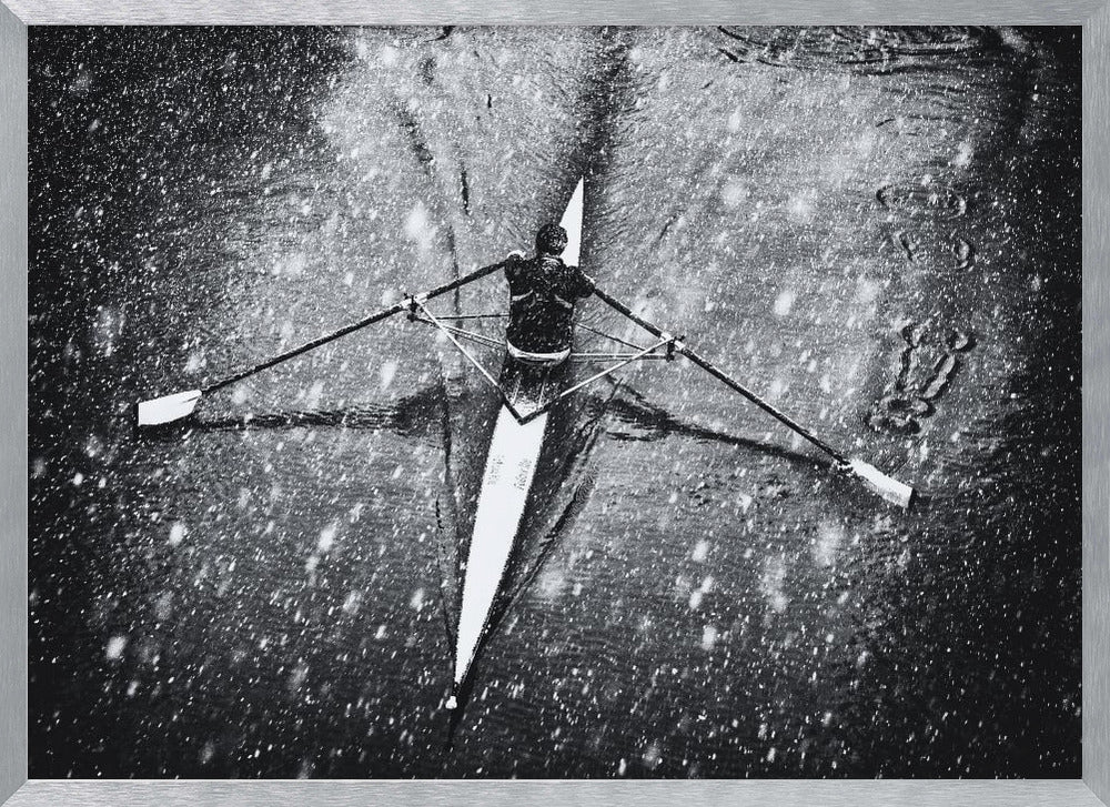 A dramatic black and white overhead photograph of a single rower in a narrow scull, captured mid-stroke as they cut through dark water during a heavy snowfall, creating a high-contrast and atmospheric scene. Decor