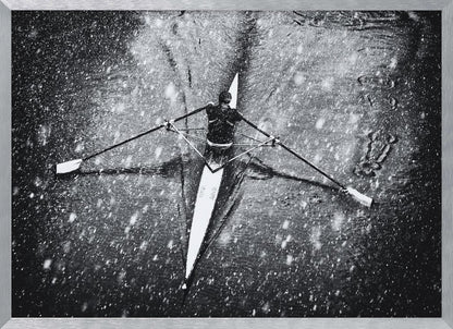 A dramatic black and white overhead photograph of a single rower in a narrow scull, captured mid-stroke as they cut through dark water during a heavy snowfall, creating a high-contrast and atmospheric scene. Decor