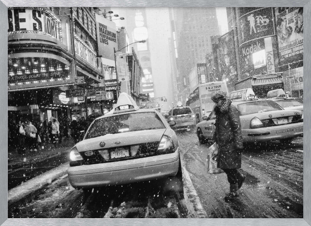 A black and white photograph of a bustling New York City street during a snowstorm. A person in a heavy winter coat crosses the street in front of a line of yellow cabs, with the bright lights of Times Square billboards glowing softly in the background through the falling snow. Print