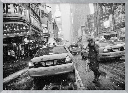 A black and white photograph of a bustling New York City street during a snowstorm. A person in a heavy winter coat crosses the street in front of a line of yellow cabs, with the bright lights of Times Square billboards glowing softly in the background through the falling snow. Print