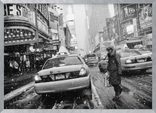 A black and white photograph of a bustling New York City street during a snowstorm. A person in a heavy winter coat crosses the street in front of a line of yellow cabs, with the bright lights of Times Square billboards glowing softly in the background through the falling snow. Print