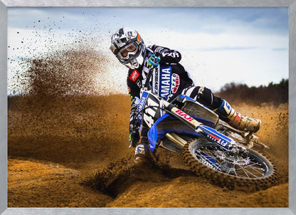 A motocross rider in full gear leans sharply into a turn on a dirt track, kicking up a large spray of mud and dirt from their blue and white Yamaha motorcycle. Artwork