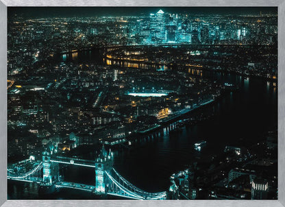 A framed aerial photograph of London at night, featuring the iconic Tower Bridge illuminated in bright teal in the foreground and the glowing skyscrapers of Canary Wharf across the River Thames in the background. Artwork