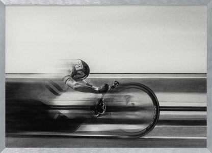 A black and white photograph captured with motion blur, showing a cyclist in an aerodynamic position racing at high speed. The background is blurred into horizontal lines, emphasizing the velocity. The image is enclosed in a silver frame. Wall Art