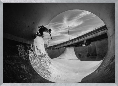 A dynamic black and white action shot of a skateboarder mid-trick on the edge of a concrete ramp in a graffiti-covered skatepark, framed by the curve of a large pipe. Print
