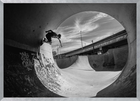 A dynamic black and white action shot of a skateboarder mid-trick on the edge of a concrete ramp in a graffiti-covered skatepark, framed by the curve of a large pipe. Print