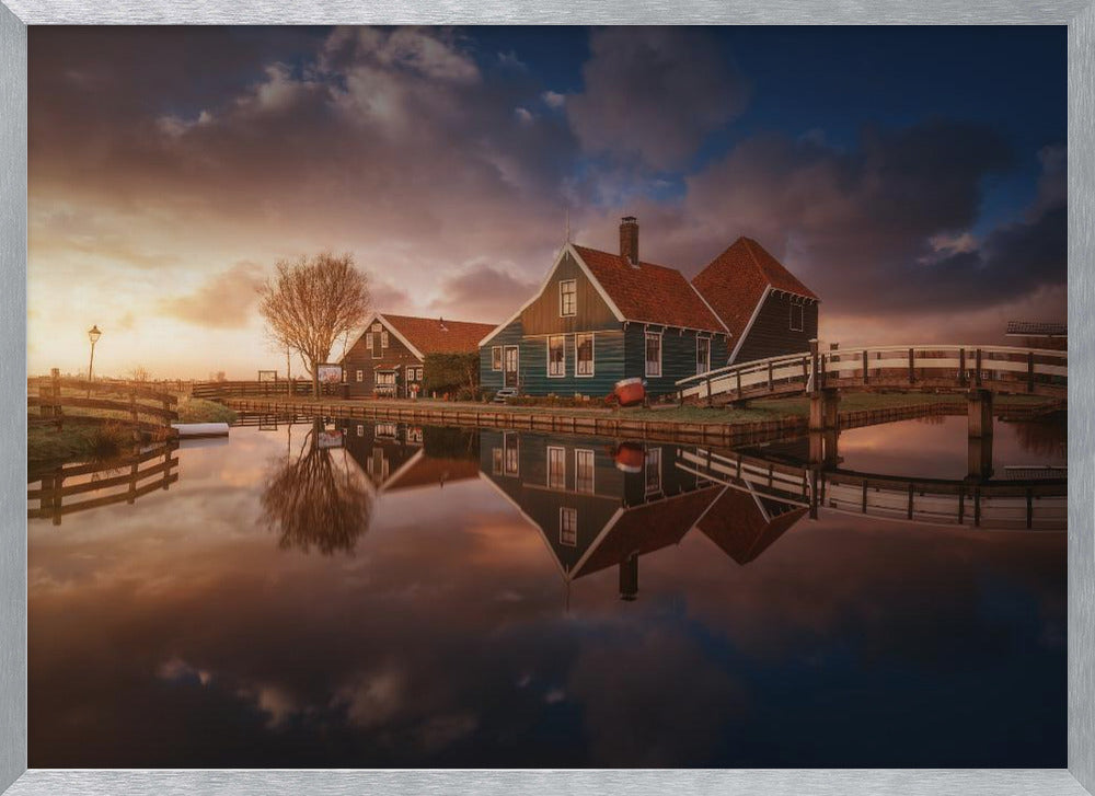 A picturesque landscape of traditional Dutch houses with teal walls and red roofs reflected perfectly in the calm water of a canal at sunset, under a dramatic, cloudy sky. A small wooden bridge crosses the canal, and the scene is enclosed in a silver frame. Artwork